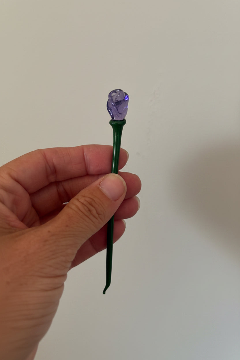 Hand holding a hairpin with a purple flower head against a white background