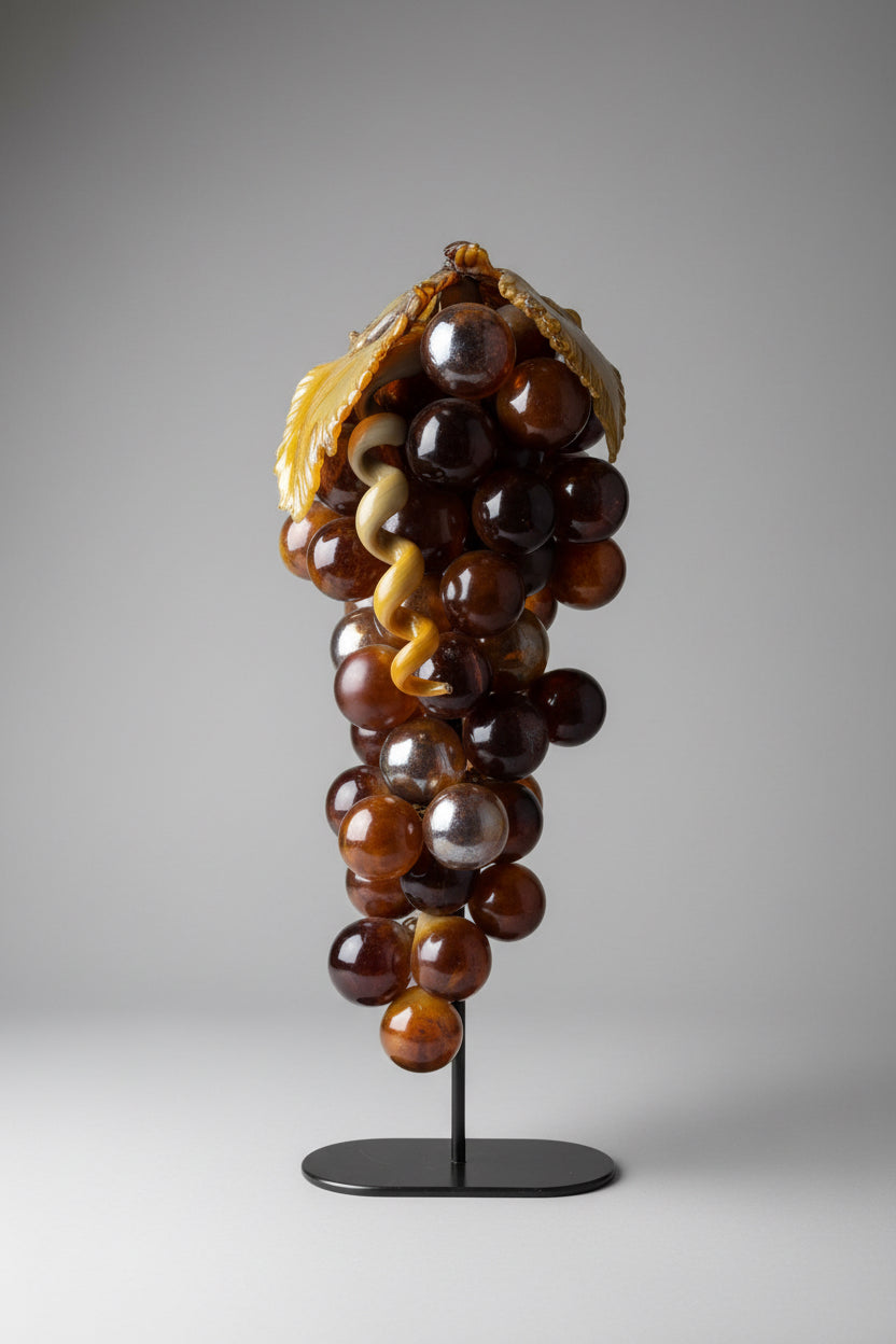 A decorative sculpture of glossy dark brown glass grapes on a stem, mounted on a black display stand against a textured off-white wall.