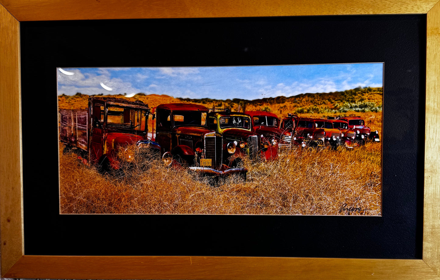 A framed landscape art print depicting vintage red trucks in a dry field under a blue sky, mounted on a textured white wall with a black mat and a light wood frame.