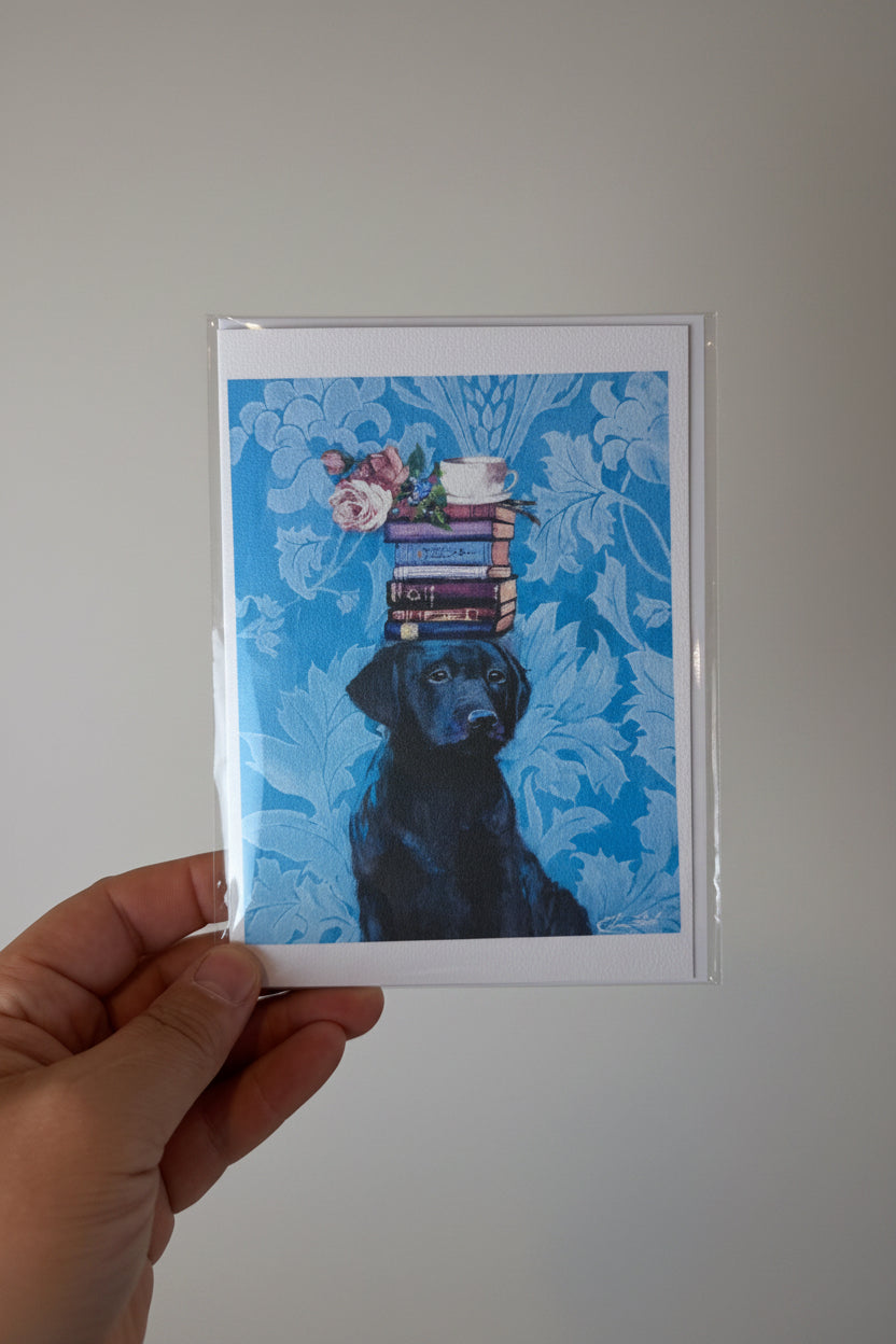 A hand holding a square art print featuring a black dog balancing a stack of books and flowers against a bright blue patterned background, shown against a textured off-white wall.