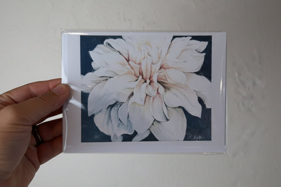 A hand holding a clear plastic-wrapped art print featuring a large white flower with soft pink highlights against a dark navy background, set against a textured off-white wall.