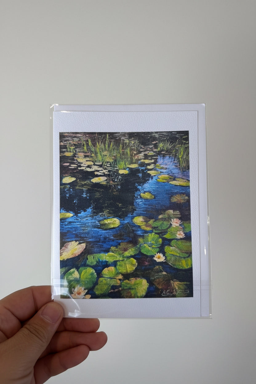 A hand holding a small art print or card featuring a pond scene with green lily pads and blue water, photographed against a textured white wall.