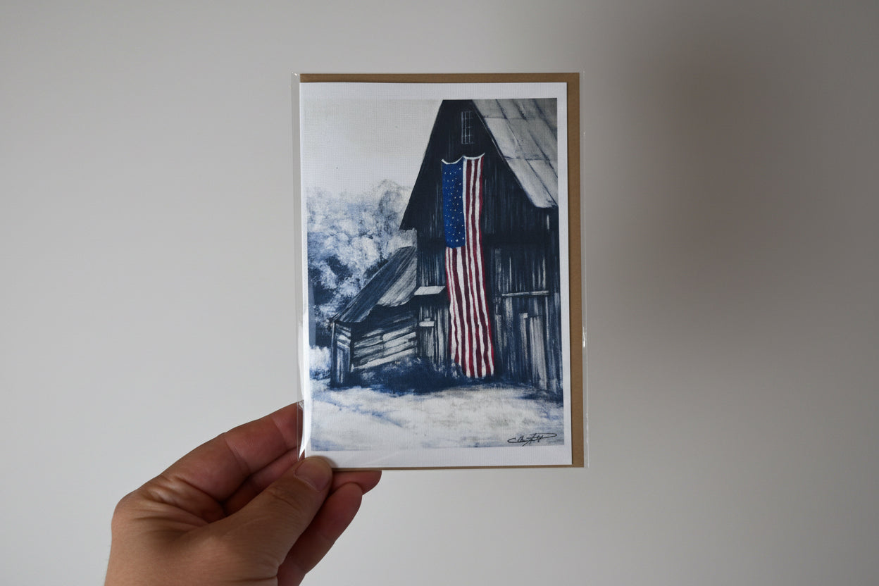 A hand holds a greeting card featuring a watercolor-style illustration of a rustic barn with an American flag hanging in front, against a textured white wall.