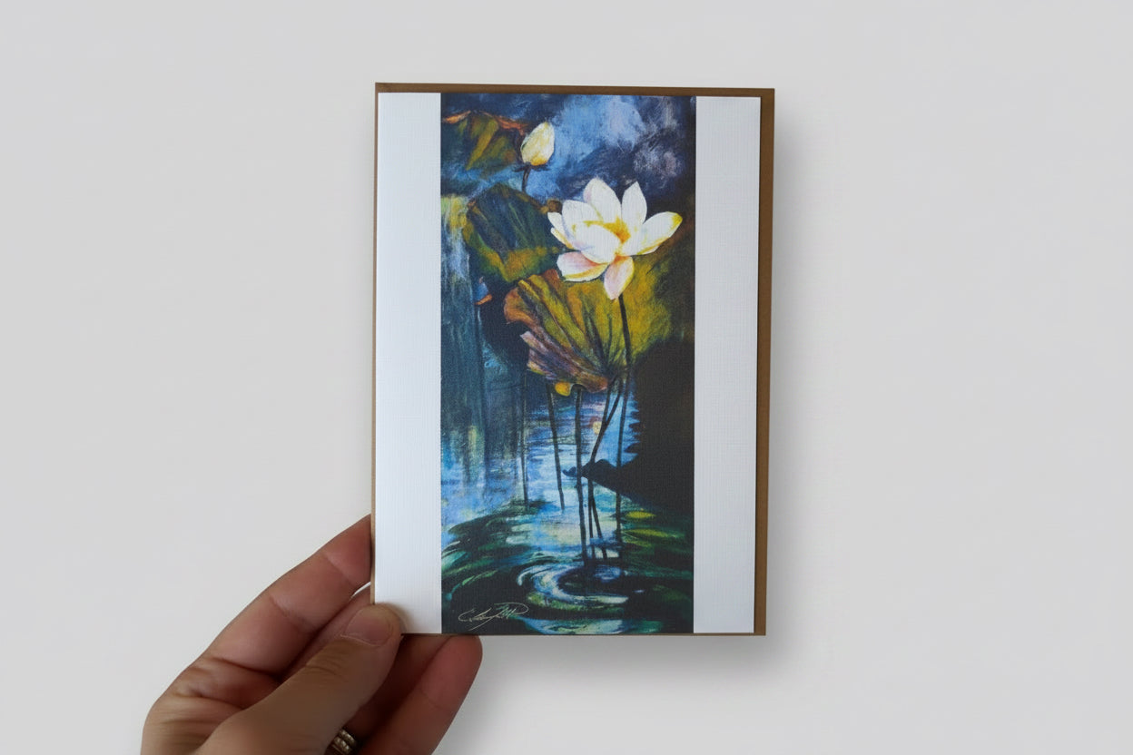 A hand holding a square art print or card featuring a painted water lily scene with white flowers, green and yellow lily pads, and blue water, against a textured white wall.