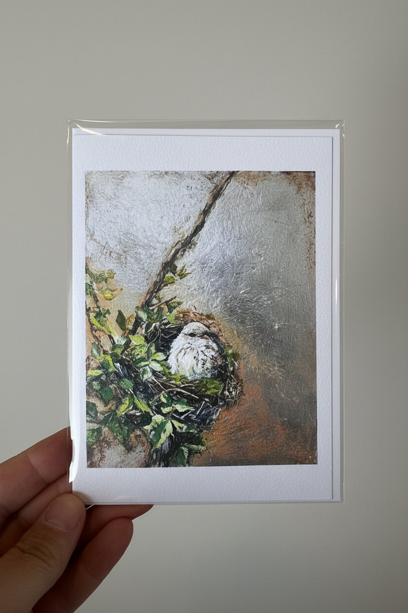 A hand holding a matted art print featuring a textured painting of a bird's nest with green leaves, photographed against a white textured wall.