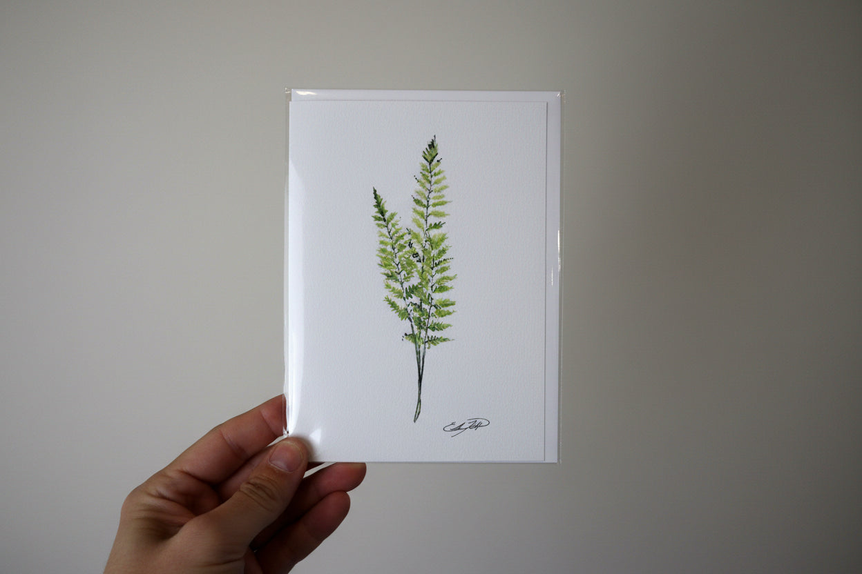 A hand holding a square white art print or card featuring a green fern illustration, photographed against a textured off-white wall.