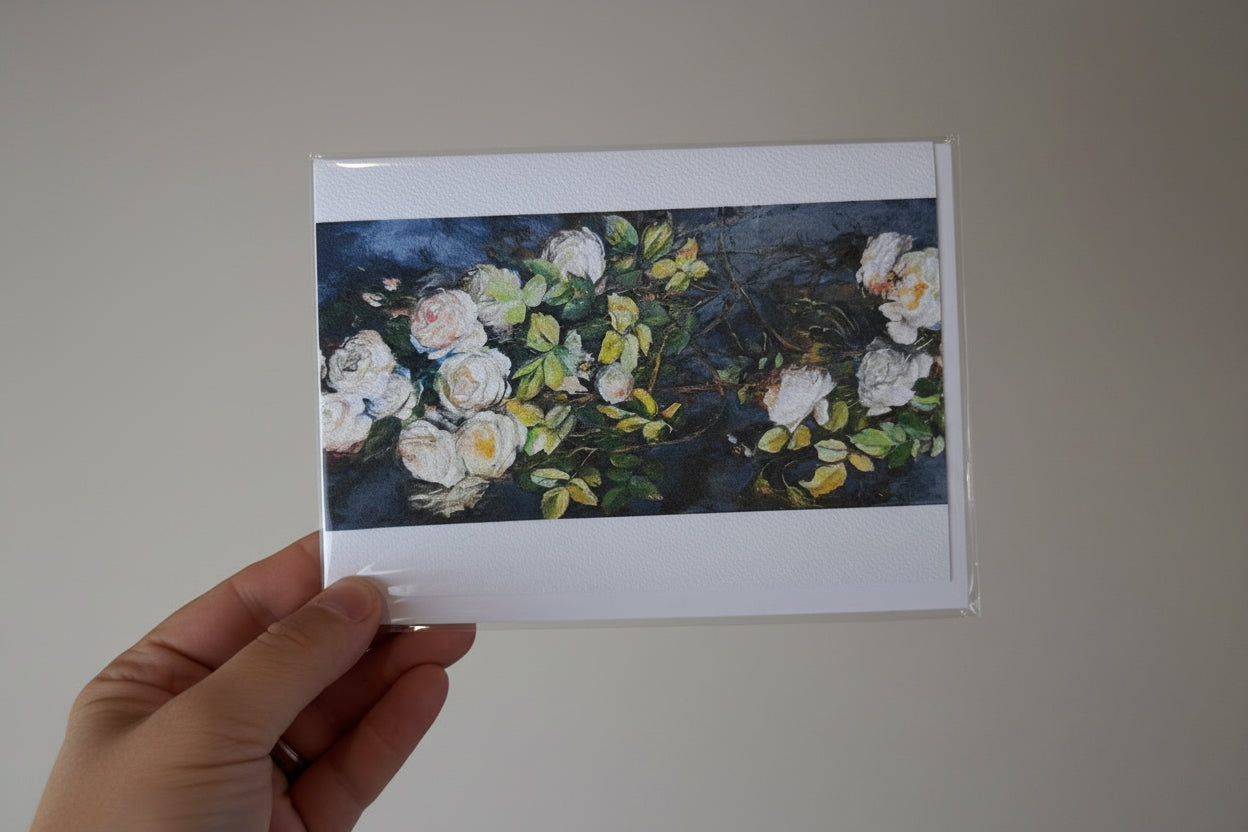 A hand holding a printed card or art print featuring a bouquet of white flowers with green leaves on a dark blue background, photographed against a textured white wall.